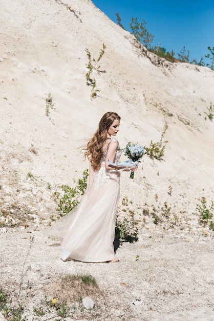 Beautiful brunette bride with flying curls on the background of a sandy mountain in a stylish dress and a blue and white bouquet.