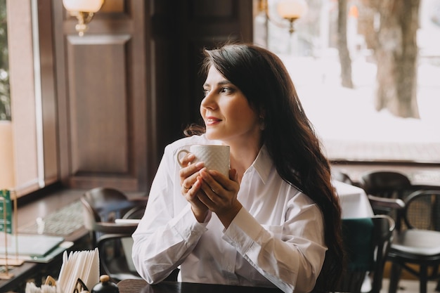 카페에서 자연광과 함께 흰색 컵에 커피를 마시며 웃고 있는 비즈니스 정장을 입은 아름다운 브루네트