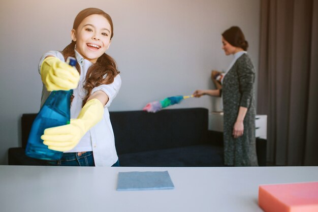 Beautiful brunette caucasian mother and daughter cleaning together in room. Nice funny girl look on caera and use psray on it. She plays. Her mom blast the dust behind.