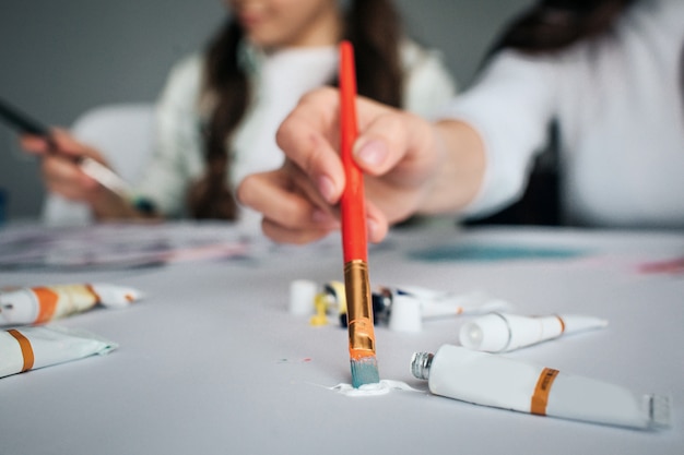 Beautiful brunette caucasian mother and daughter paint together in room. Close up of hand holding brush and put in into white paint. Cut view. Tubes with paint.