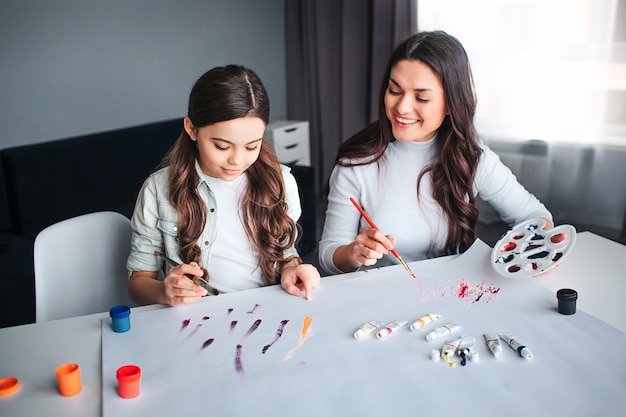 Beautiful brunette caucasian mother and daughter paint together in room. Young woman look at girl and smile. She hold palette. Child painting concentrated.