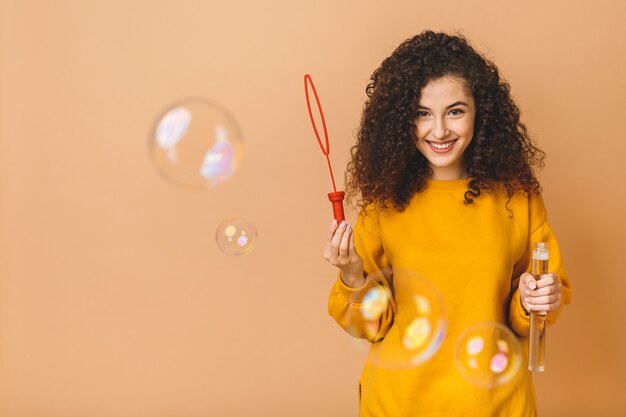 La bella ragazza riccia castana dello studente indossa le bolle di sapone di fabbricazione casuali isolate su fondo beige.