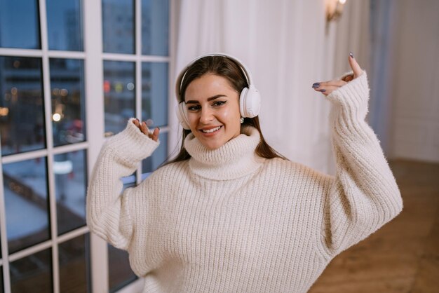 Beautiful brunette female model in white sweater and headphones dancing home against window