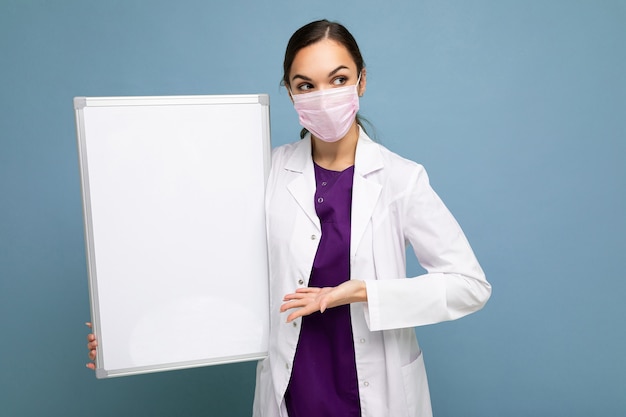 Beautiful brunette female nurse in protective face mask and white medical coat holding an empty