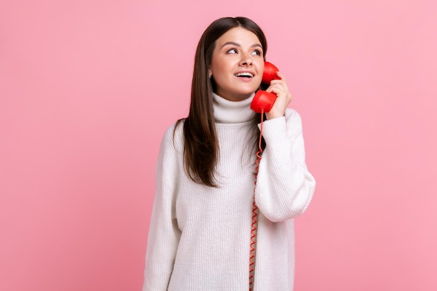 Beautiful brunette female with dreamy expression talking on red retro telephone, holding handset, wearing white casual style sweater. Indoor studio shot isolated on pink background.