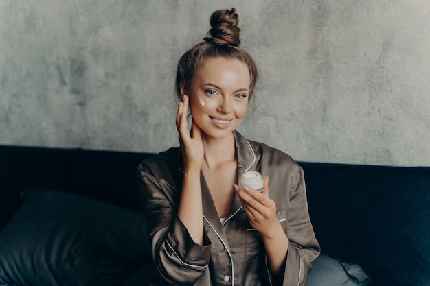 Bella donna bruna con pelle sana si prepara per il trucco mattutino, applicando la crema per il viso mentre è seduta sul letto a casa, guardando la telecamera con un sorriso morbido sul viso. concetto di bellezza