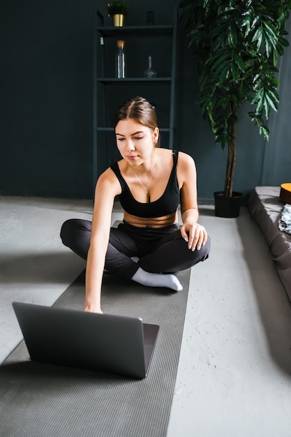 美しいブルネットのフィットネス女性は、自宅で屋内でヨガをしながら、フロントラップトップでストレッチ運動をします