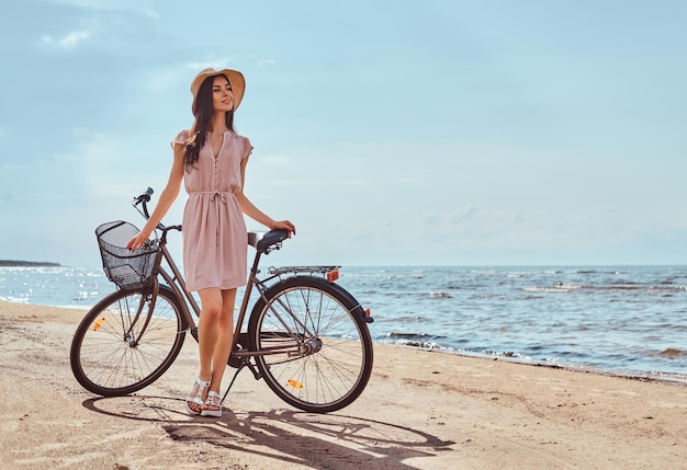 Beautiful brunette girl dressed in dress and hat posing with a bicycle on the beach on a sunny day