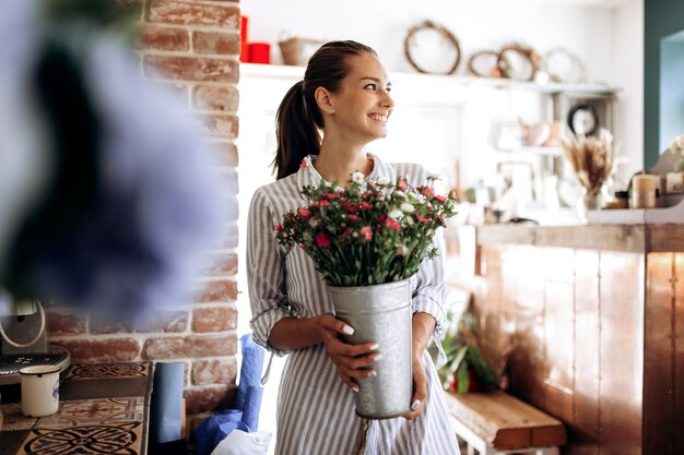 Beautiful brunette girl dressed in a striped dress holds a vase with pink and white chrysanthemums in the flower shop .