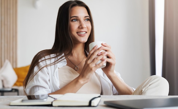 Foto bella ragazza bruna è seduta al tavolo davanti al computer portatile, tenendo la tazza di caffè con le mani.