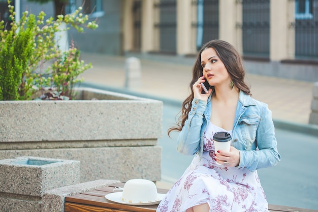 Красивая брюнетка девушка в джинсовой куртке разговаривает по телефону, сидя на скамейке в городе и проведение кофе