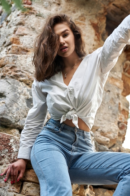 Bella ragazza bruna in jeans e una camicia bianca in posa sulle rovine nella foresta