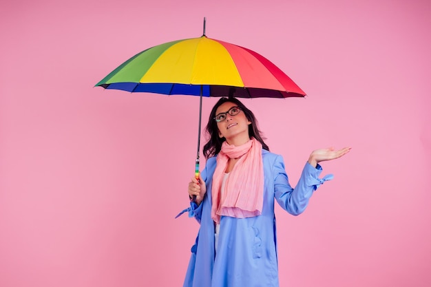 Beautiful brunette girl in the studio on a pink background in the style blue raincoat and chiffon scarf with rainbow an umbrella autumn fashion look seasonal spring sale