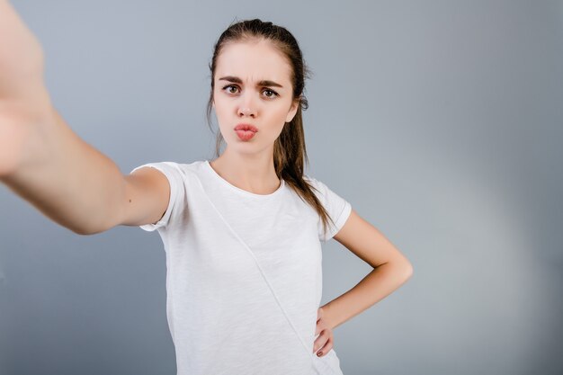 灰色で分離されたselfieを作る白いシャツを着て美しいブルネットの少女