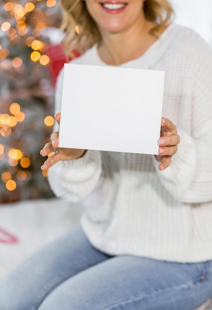 A beautiful brunette girl in a white sweater sits on a bed
against the background of christmas lights and holds a white card
in her hands free space for text