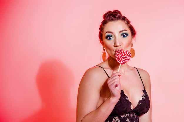 Photo beautiful brunette girl with blue makeup, pigtails and a sexy black top posing against a red background with a heart-shaped lollipop. horizontal photo. copy space