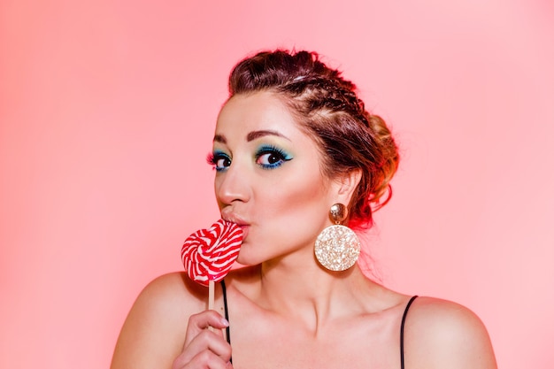 Beautiful brunette girl with blue makeup, pigtails and a sexy black top posing against a red background with a heart-shaped lollipop. Horizontal photo