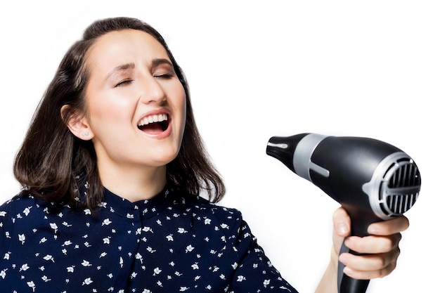 Beautiful brunette girl with eyes closed screaming singing into a Hairdryer on hand isolate the blue shirt happiness emotions the joy of fooling around