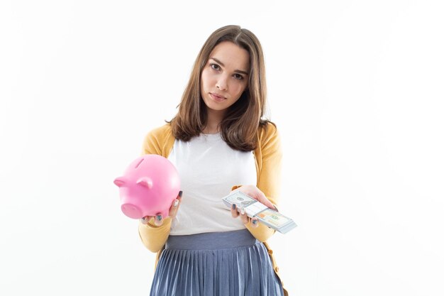 A beautiful brunette holds a pink piggy bank in one hand and a wad of money in the other. Concept of depositing money in a bank