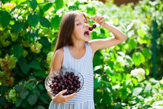 写真 美しい茶色の小さな女の子が庭でチェリーのボウルを握っている