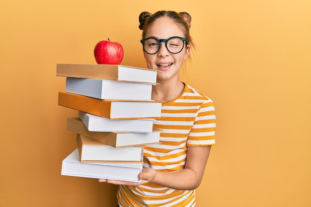 Photo beautiful brunette little girl holding a pile of books with an apple on the top winking looking at the camera with sexy expression, cheerful and happy face.