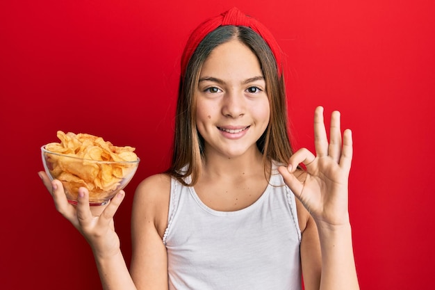 감자 칩을 들고 있는 아름다운 갈색 머리 소녀 손가락으로 확인 표시를 하고 친절한 몸짓으로 훌륭한 기호를 미소 짓고 있습니다.