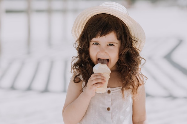 Beautiful brunette little girl in white dress and hat eating ice cream on beach in summertime