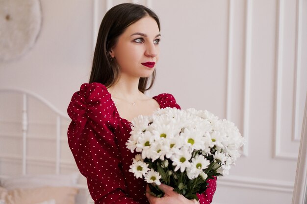 Beautiful brunette in a red dress with a bouquet of daisies at home