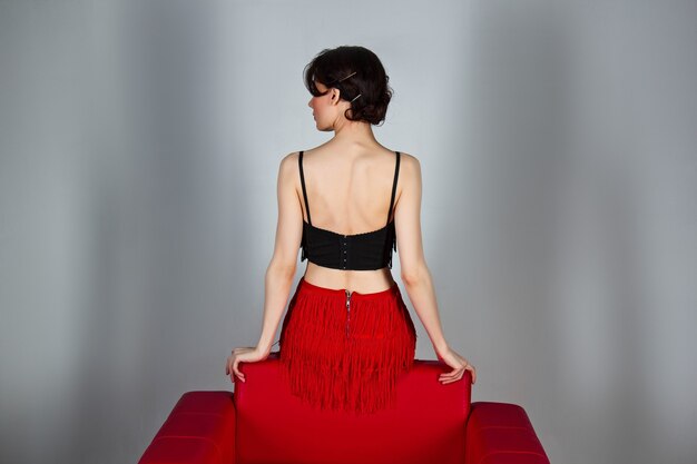 a beautiful brunette in red shoes sits in a red armchair on a gray background.