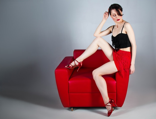 a beautiful brunette in red shoes sits in a red armchair on a gray background.