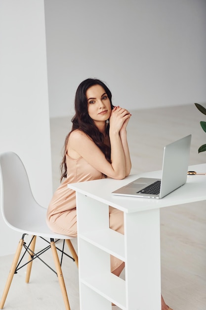 Beautiful brunette with curly hair in luxury clothes sits by table with laptop at home at daytime