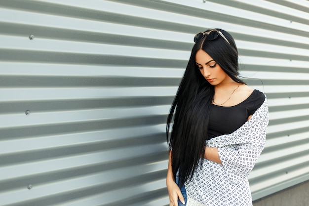 Photo beautiful brunette with long hair in fashionable clothes near a metal wall in the street