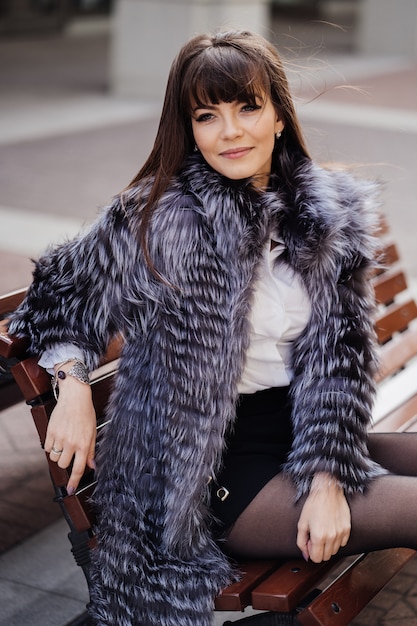 Beautiful brunette with the long straight hair wearing in a grey fur coat, white shirt and black skirt siting  on the bench and smiles thoughtfully