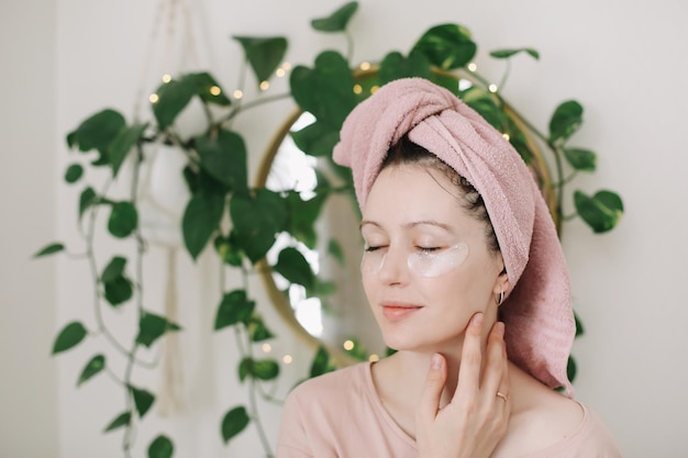 Beautiful brunette with towel on her head using eye patches Morning habits and home beauty routine concept