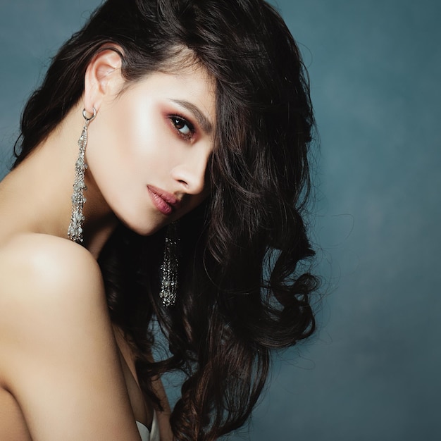 Красивая брюнетка женщина крупным планом портрет молодая идеальная женщина модель лицо на синем фоне длинные здоровые волнистые волосы макияж серебро и полудрагоценные камни серьги