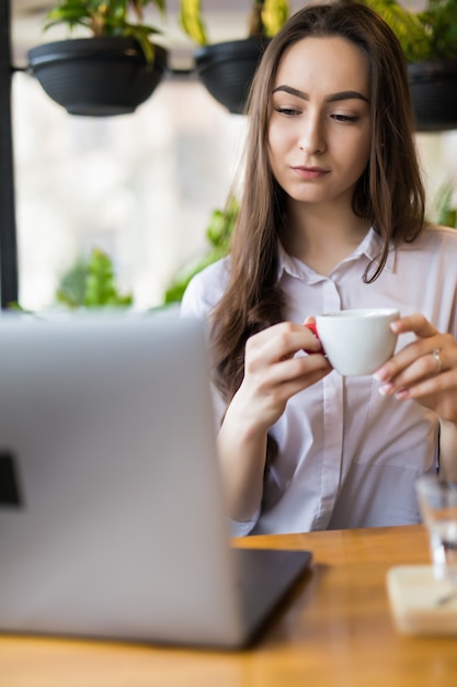 Beautiful brunette woman drinking coffee and using laptop in cafe