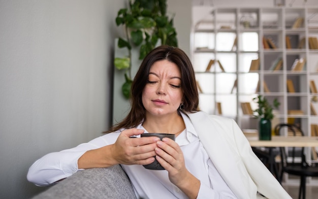 A beautiful brunette woman drinks tea in a mug on the sofa in the room. Copy of the space