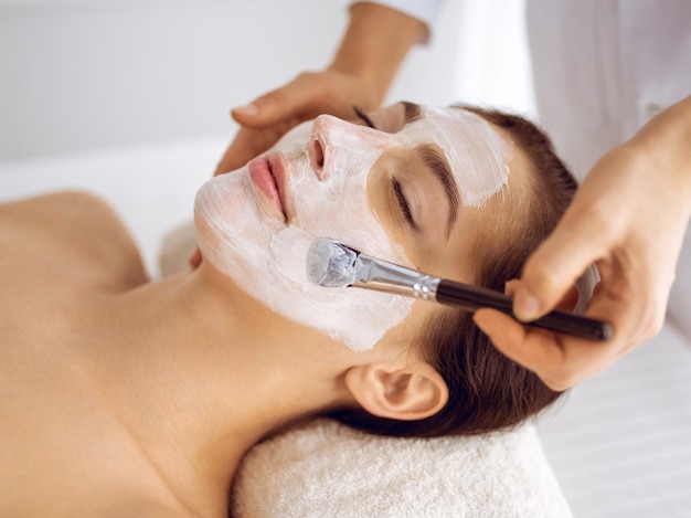 Beautiful brunette woman enjoying applying cosmetic mask with closed eyes. Relaxing treatment in medicine and spa center concepts.