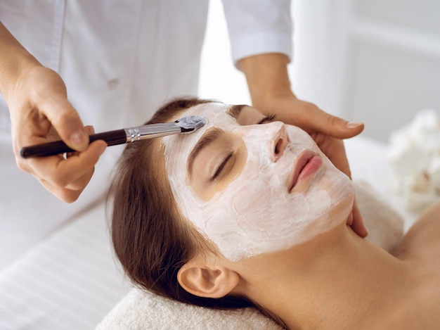 Bella donna castana che si diverte ad applicare la maschera cosmetica con gli occhi chiusi. trattamento rilassante nei concetti di medicina e centro termale.