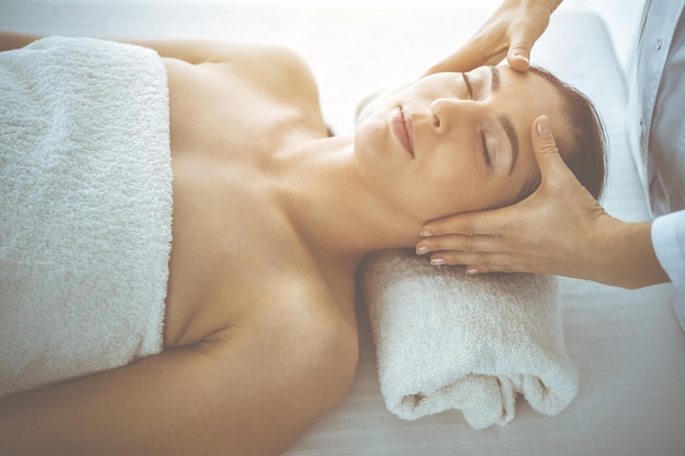 Beautiful brunette woman enjoying facial massage with closed eyes in sunny spa center. Relaxing treatment and cosmetic medicine concepts