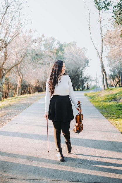 Foto bella donna castana che tiene un violino e un arco, camminando attraverso la strada del parco al tramonto.