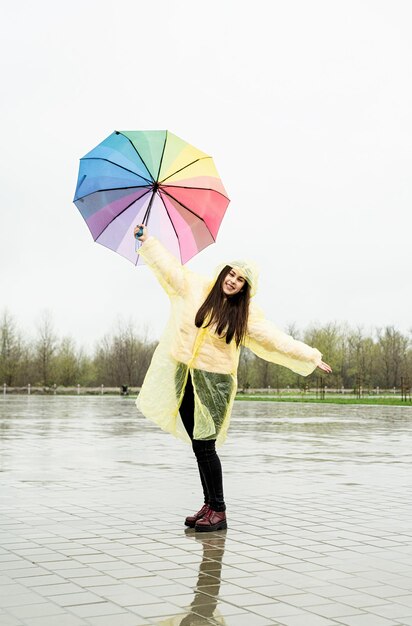 写真 黄色いレインコートを着た美しい茶色の女性が雨の中に虹の傘を握っている