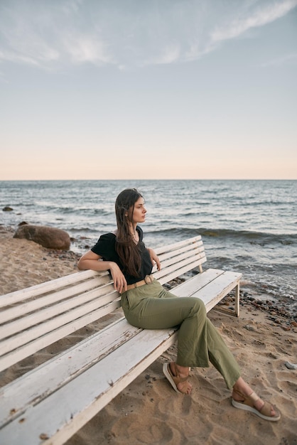 Beautiful brunette woman looking far into the sea horizon Girls sits on white bench on the beach Concept of worklife balance and wellbeing