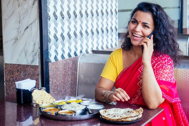 Красивая брюнетка в красном сари с аппетитом ест традиционный тхали с рискурддалом в ресторане Гоа, чай масала, она разговаривает по телефону с белой улыбкой