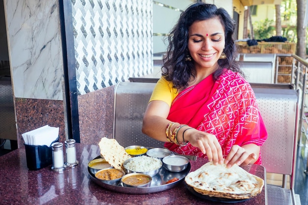 Beautiful brunette woman in red sari eating with appetite traditional thali wirh risecurddal in Goa restaurant masala tea