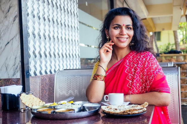 Beautiful brunette woman in red sari eating with appetite traditional thali with risecurddal in Goa restaurant and drinking masala tea