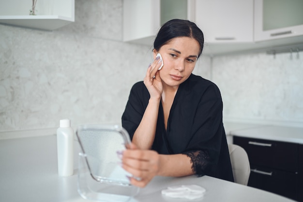 Bella donna castana che rimuove trucco dal suo fronte con lo specchio. fronte di pulizia della bella ragazza naturale carina bellezza con spugna di cotone. cosmetologia e spa, problemi di cura della pelle, concetto di trattamento dell'acne