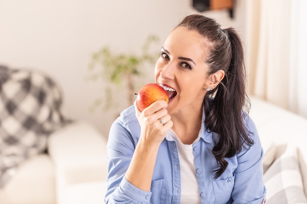 Beautiful brunette woman sitting in a living room biting off an apple.