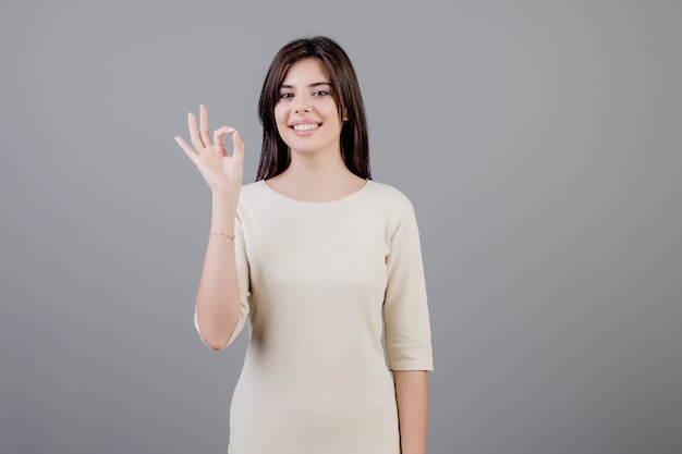 Beautiful brunette woman smiling and showing ok gesture isolated over grey