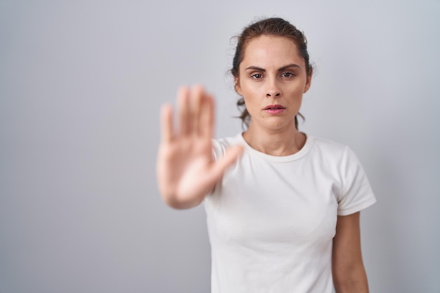 beautiful brunette woman standing over isolated background doing stop sing with palm of the hand. warning expression with negative and serious gesture on the face.